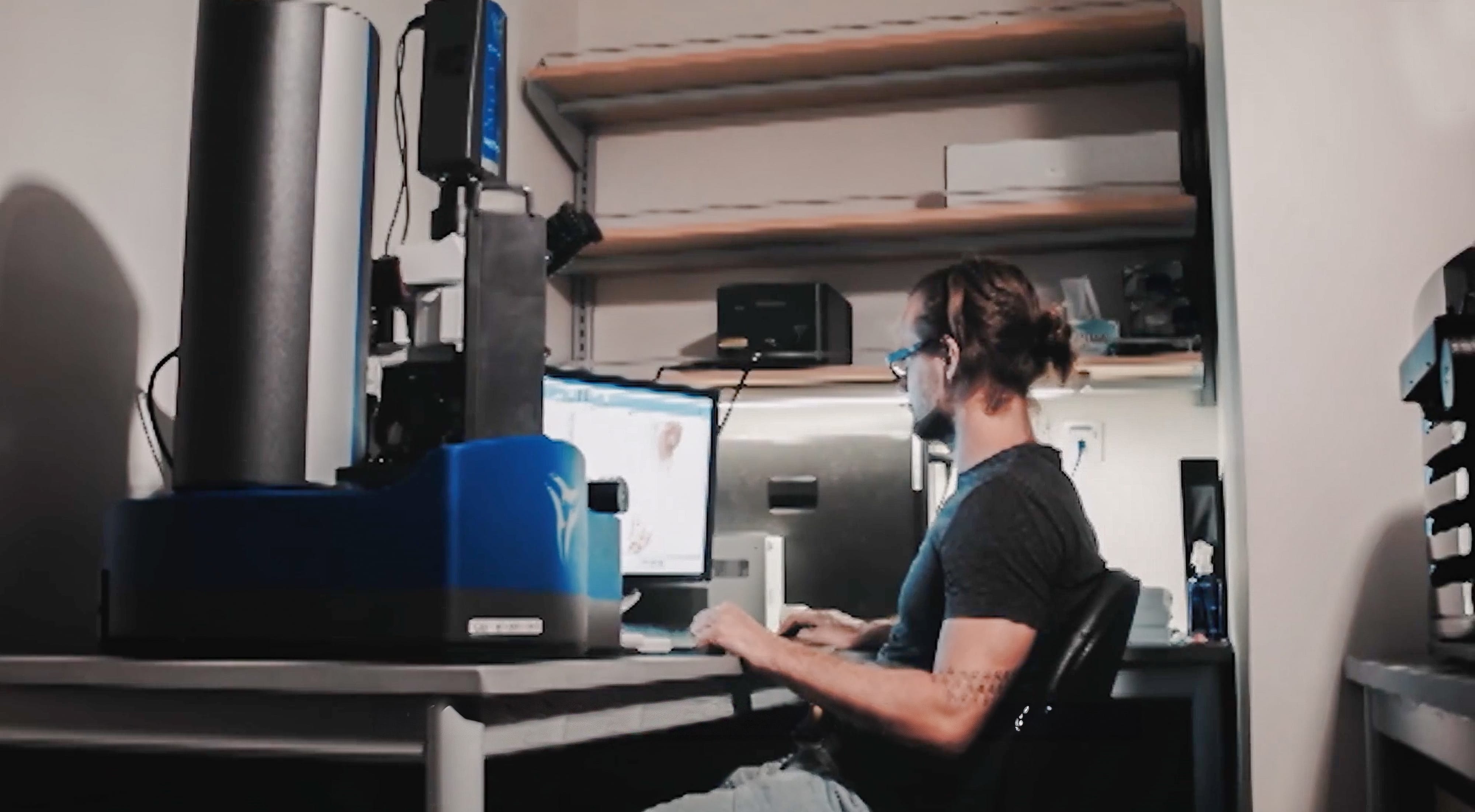 A person sits at a desk with a large computer setup, surrounded by shelves holding various equipment and supplies.